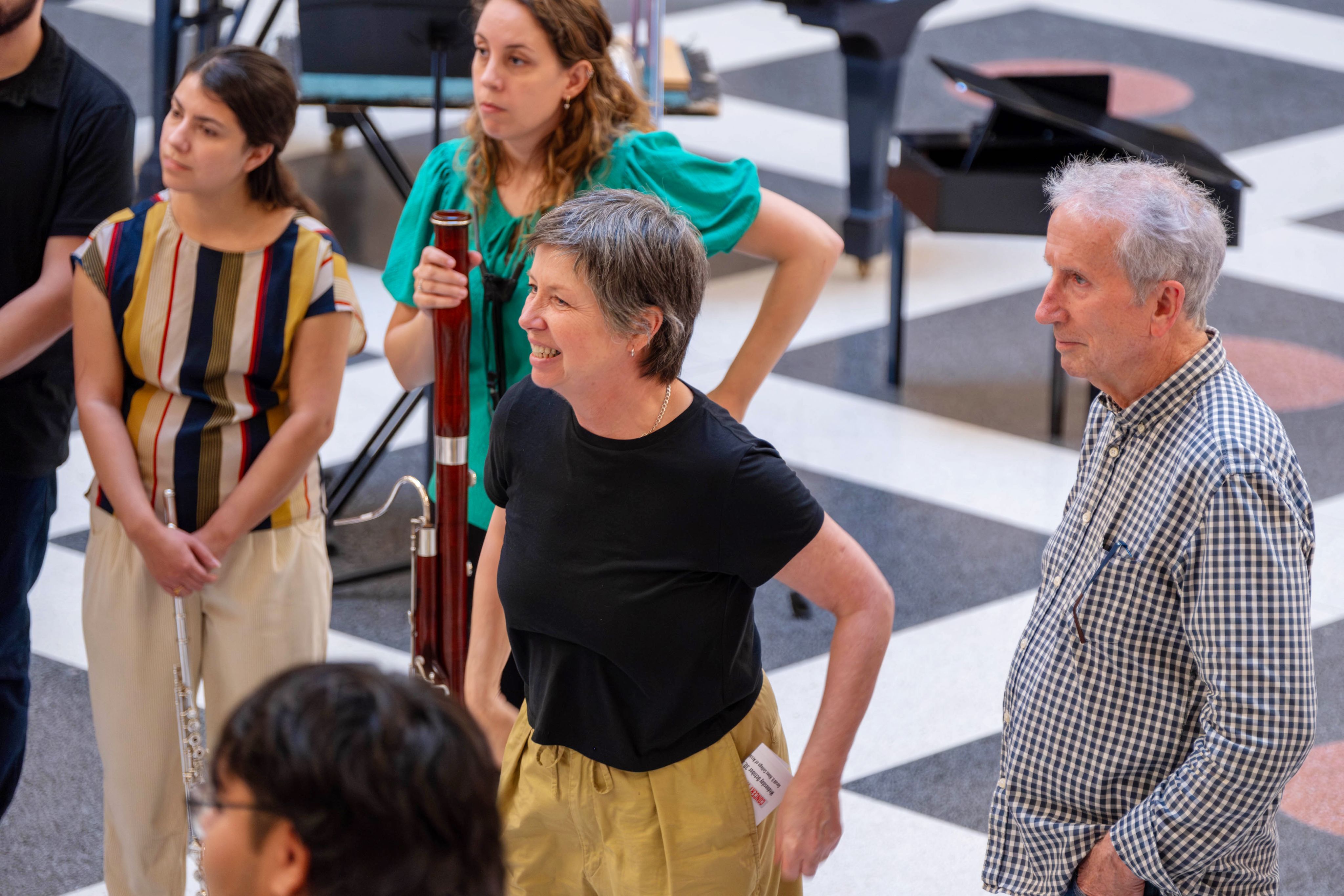 A woman and a man, presumably in their 70s, look at a group of college students intently, some holding instruments. They stand on a patterend tile floor in the architecture building at the University of Houston
