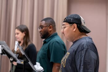 A group of three people standing next to each other in front of a microphone. The person in the front is wearing a black t-shirt and a cap, while the person to the left is holding a paper in their hand. In the background, there is a curtain and a wall.